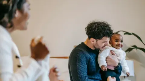 A man is holding a smiling baby while a woman looks on. 